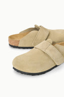 A pair of beige suede clogs with a hook-and-loop strap closure and a black outsole, shown on a clean white background.