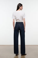 A model shown from the back wearing dark blue wide-leg jeans, a white t-shirt, and dark brown shoes, standing against a plain white studio wall.