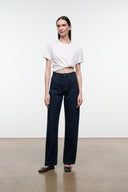 A model stands in a minimalist studio setting wearing dark blue straight-leg jeans, a white cropped t-shirt, and dark brown shoes.