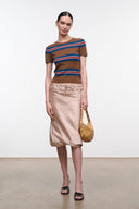 A model stands in a studio wearing a brown short-sleeve knit top with blue and red horizontal stripes, paired with a light beige midi skirt and holding a tan shoulder bag.