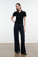 A woman modeling a black short-sleeve polo shirt paired with dark wide-leg jeans and black flat shoes, standing against a plain white studio wall.