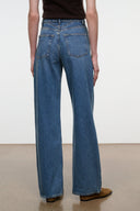 Rear view of a person wearing blue wide-leg jeans with a dark brown top and brown shoes, standing against a plain light wall and beige floor.