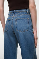 Close-up back view of a person wearing blue denim jeans with a dark brown top, showing the back pockets and waistband details against a plain light background.