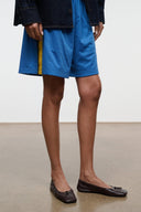 Espresso brown ballet flats with bow detail and stitched trim, worn with blue shorts.