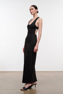 A woman modeling a floor-length black sleeveless gown with a deep scoop neckline and side cutouts, standing against a plain light studio background.