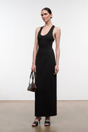 A woman modeling a sleeveless black maxi dress with a scoop neckline, paired with black heeled sandals and a small brown handbag, standing against a plain light gray studio background.