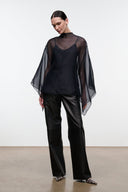 A model wearing a black sheer mesh cape-style top over a black camisole, paired with black wide-leg trousers and patterned pointed shoes, set against a minimalist studio background.