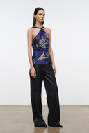 A model stands in a studio wearing a halter-neck top with a blue, gold, and white bird pattern, paired with black wide-leg pants.