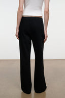 A model shown from the back wearing black wide-leg trousers and a white ribbed top, standing against a minimalist studio background with a white wall and beige floor.