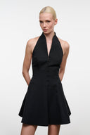 A blonde model wearing a black halter-neck mini dress with a button-front bodice and flared skirt, photographed against a plain light gray studio background.