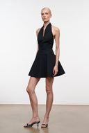 A model wearing a black halter-neck mini dress with a button front and flared skirt, paired with black and white patterned mule heels, standing against a plain white studio background.