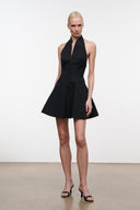 A model wearing a black halter-neck mini dress with a button-front bodice and flared skirt, paired with black strappy heels, standing against a plain white studio background.