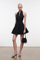 A model stands in a studio wearing a black halter-neck mini dress with a flared skirt and button front, holding a black feather-trimmed handbag and wearing black strappy heels.