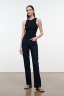 A woman modeling a black sleeveless ribbed tank top and dark blue straight-leg jeans with pink sandals, standing against a plain white studio wall.