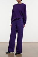 A model wearing a matching deep purple knit crewneck sweater and wide-leg trousers, standing against a plain off-white studio background.
