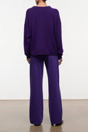 A model shown from the back wearing a matching purple knit lounge set, consisting of a crewneck sweatshirt and wide-leg trousers, against a plain white wall.