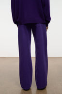 A person shown from the back wearing a matching deep purple sweatshirt and wide-leg trousers, standing against a plain white wall on a concrete floor.