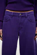 Close-up of a person wearing purple wide-leg jeans and a matching purple sweater, showing the front waistband, button, pockets, and hands against a plain light background.