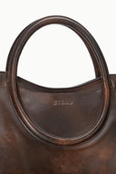 Dark brown distressed leather satchel close-up: oval handle edge and 'STAUD' logo
